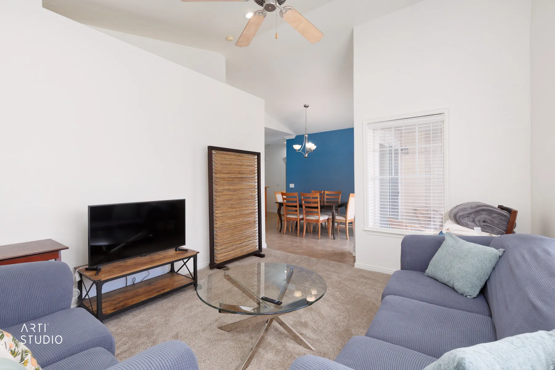 Vaulted ceiling, ceiling fan, window with blinds, carpeted flooring, and a visible dining area with a chandelier