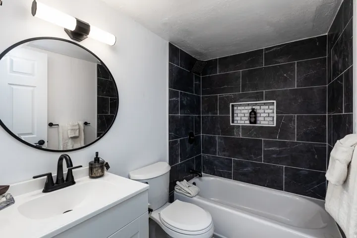 Bathroom featuring a vanity with a white countertop, a circular mirror, a toilet, a bathtub, and dark rectangular wall tiling with a recessed niche