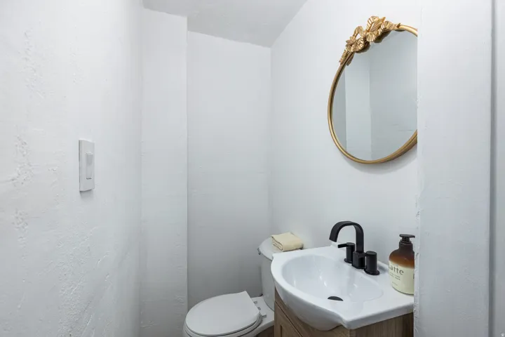 White textured walls with a built-in vanity featuring a white basin sink, black fixture, and a gold-framed circular mirror