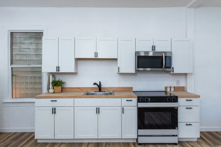 White shaker-style cabinetry with matte black hardware, wood-finish countertops, stainless steel microwave, black electric range, and wood-finish flooring