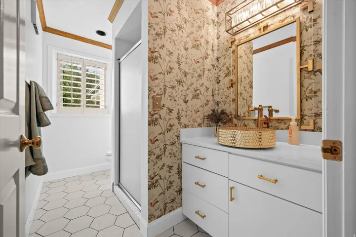 White hexagonal floor tiles, plantation shutters, custom vanity with a white countertop, gold-tone vessel sink, and a framed mirror with integrated lighting