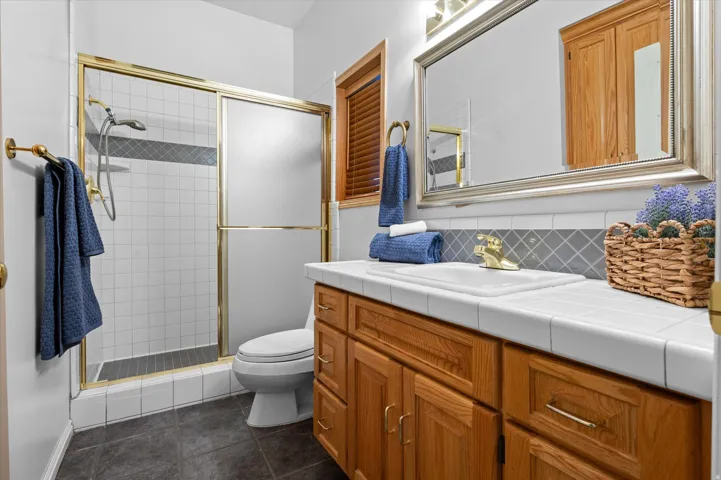 Tile flooring, wood-finish vanity with gold hardware, tiled countertop, gold-finish faucet, and a shower with sliding glass doors