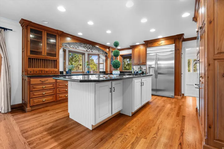 Hardwood flooring throughout, custom wood cabinetry with glass-front display, white cabinetry island with dark countertop, stainless steel refrigerator, and recessed lighting