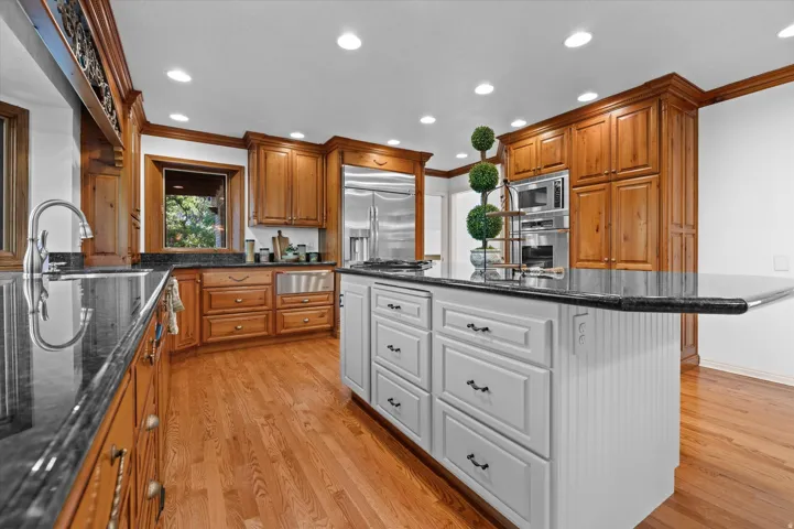 Kitchen featuring wood-finish cabinetry, stainless steel appliances, dark stone countertops, a white paneled island, double ovens, warming oven and recessed lighting