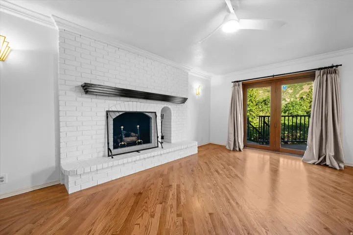 Hardwood flooring, painted brick fireplace with hearth, crown molding, double French doors, and ceiling fan with integrated lighting