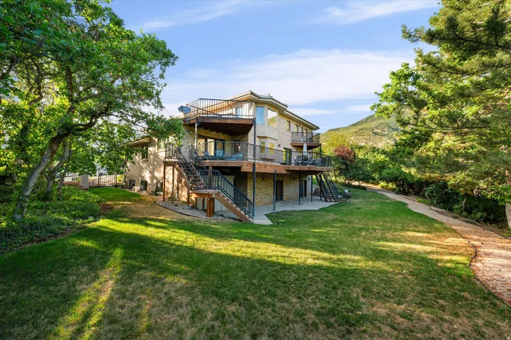 Multi-level stone exterior with multiple balconies, expansive lawn, mature trees, and a concrete patio