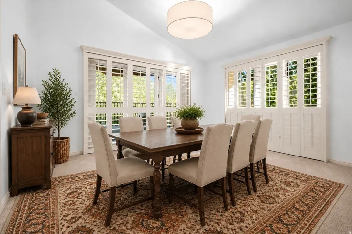Vaulted ceiling, drum pendant light fixture, window shutters, wood-finish console cabinet, and carpet flooring