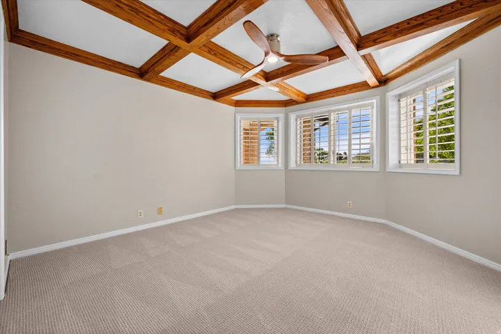 Coffered ceiling with exposed wood beams, ceiling fan, multiple windows with plantation shutters, and neutral carpeting