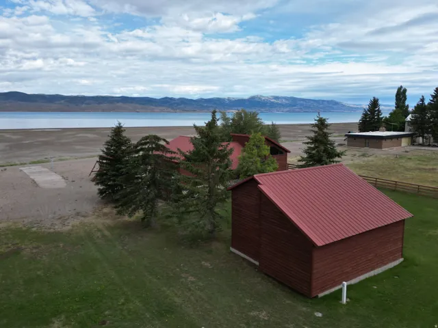 Expansive green lawn with mountain-lake backdrop, multiple structures with red metal roofs, and mature evergreen trees