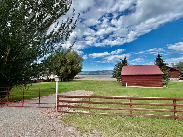 Expansive green lawn with mature trees, red metal gate entry, gravel driveway, and a red structure with a metal roof