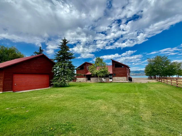 Red siding residence with stone accents, a detached red garage, lush green lawn, and a natural wood fence bordering a body of water