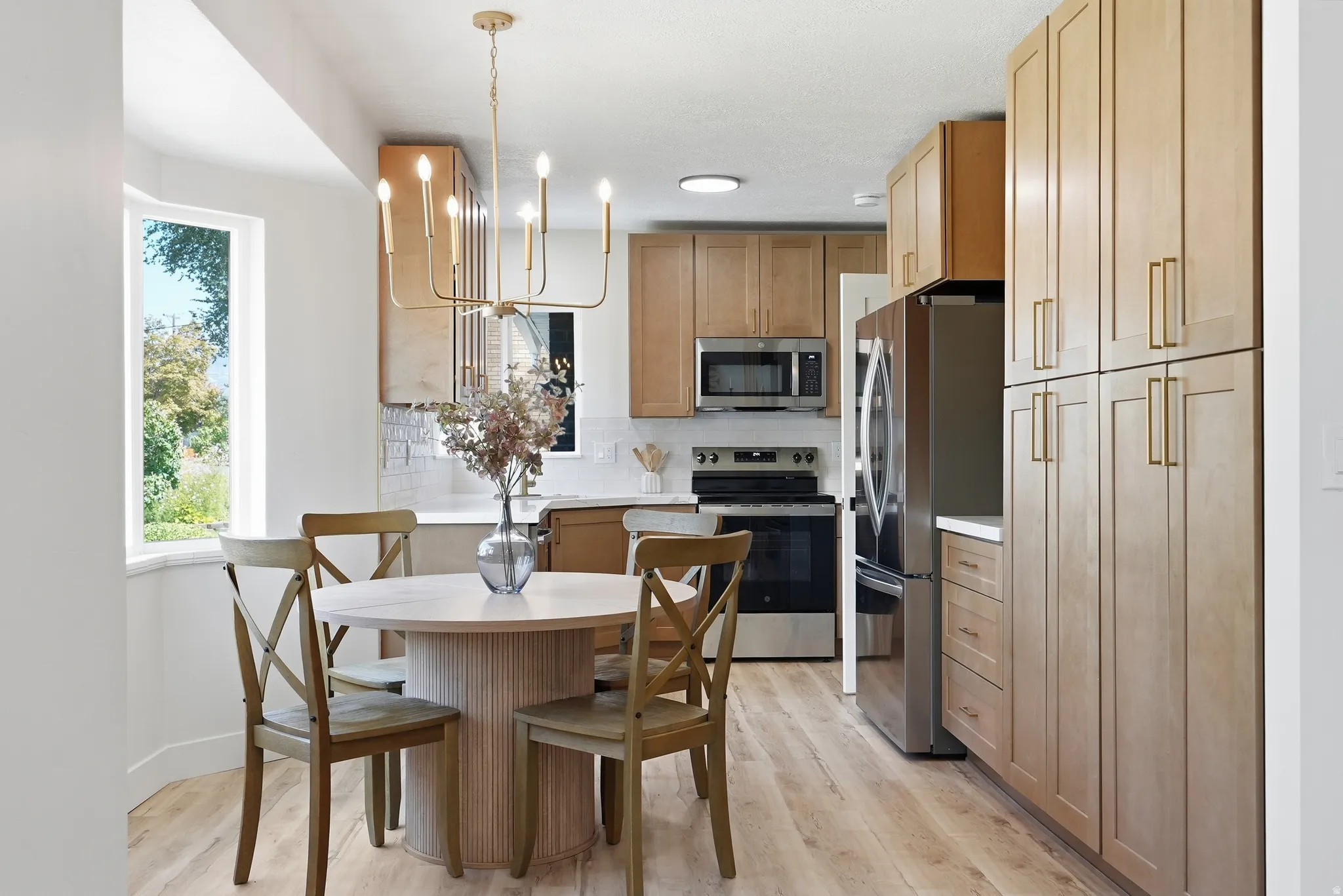 Kitchen featuring wood-finish flooring, light wood cabinetry, stainless steel appliances, a tile backsplash, and a modern chandelier