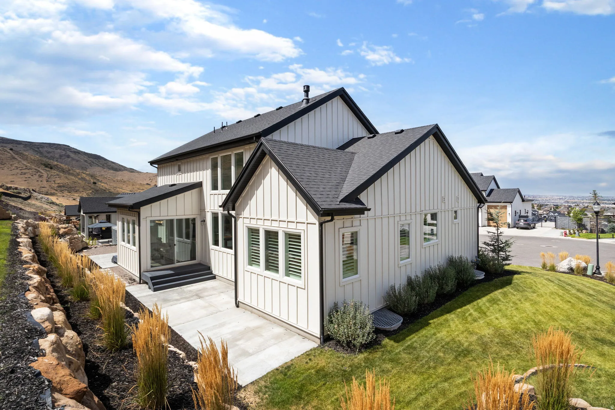 Board and batten siding, black trim accents, a dark shingled roof, a ground-level patio, and a tiered lawn