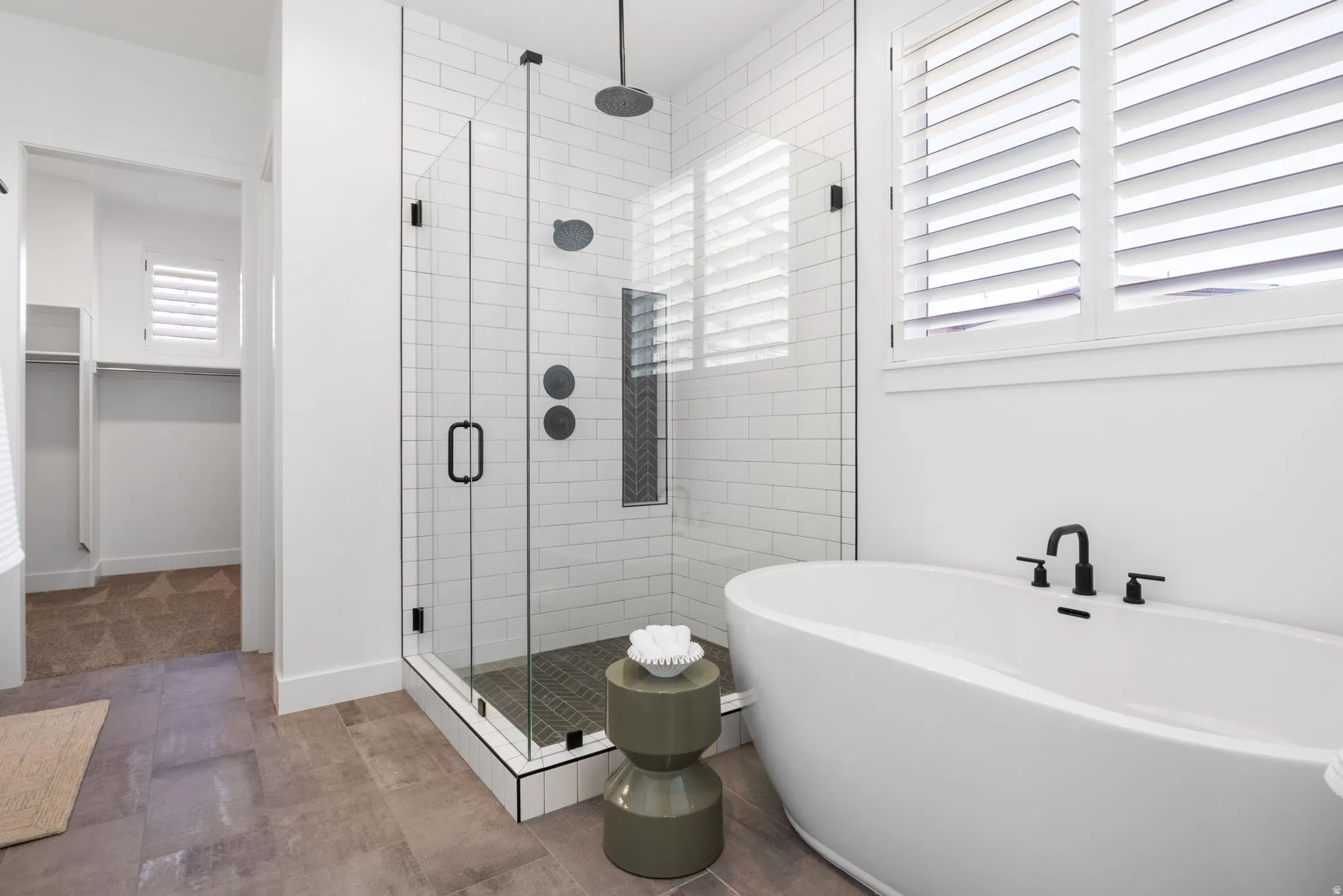 Freestanding soaking tub with matte black fixtures, glass-enclosed shower with subway tile surround, wood-finish flooring, and plantation shutters
