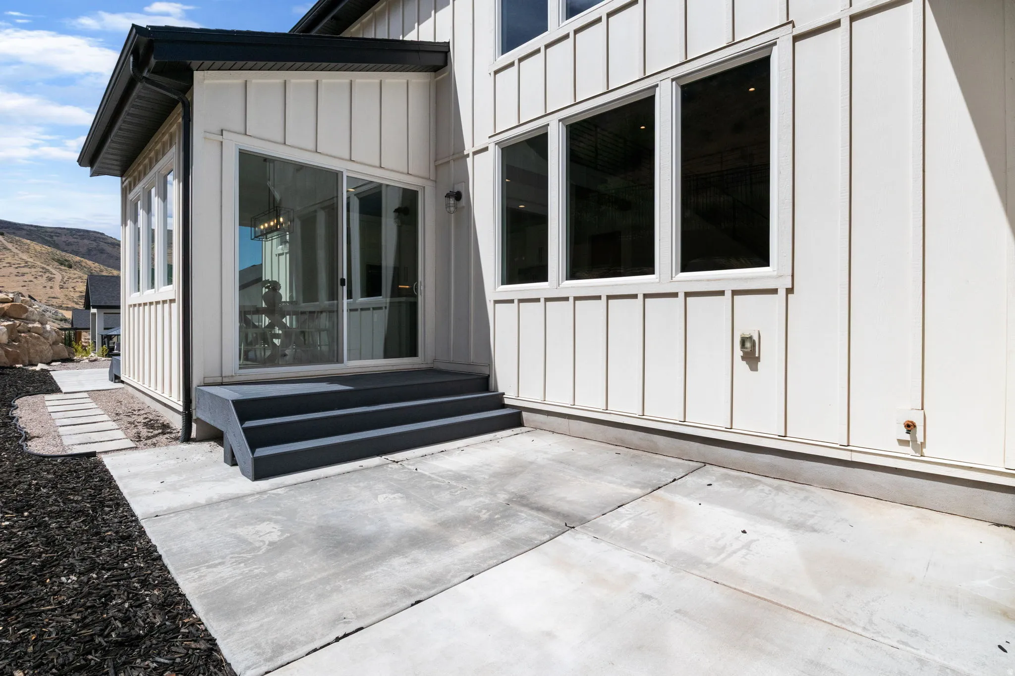 Board and batten siding, multiple large windows, dark-toned stairs, concrete patio, and a sliding glass door