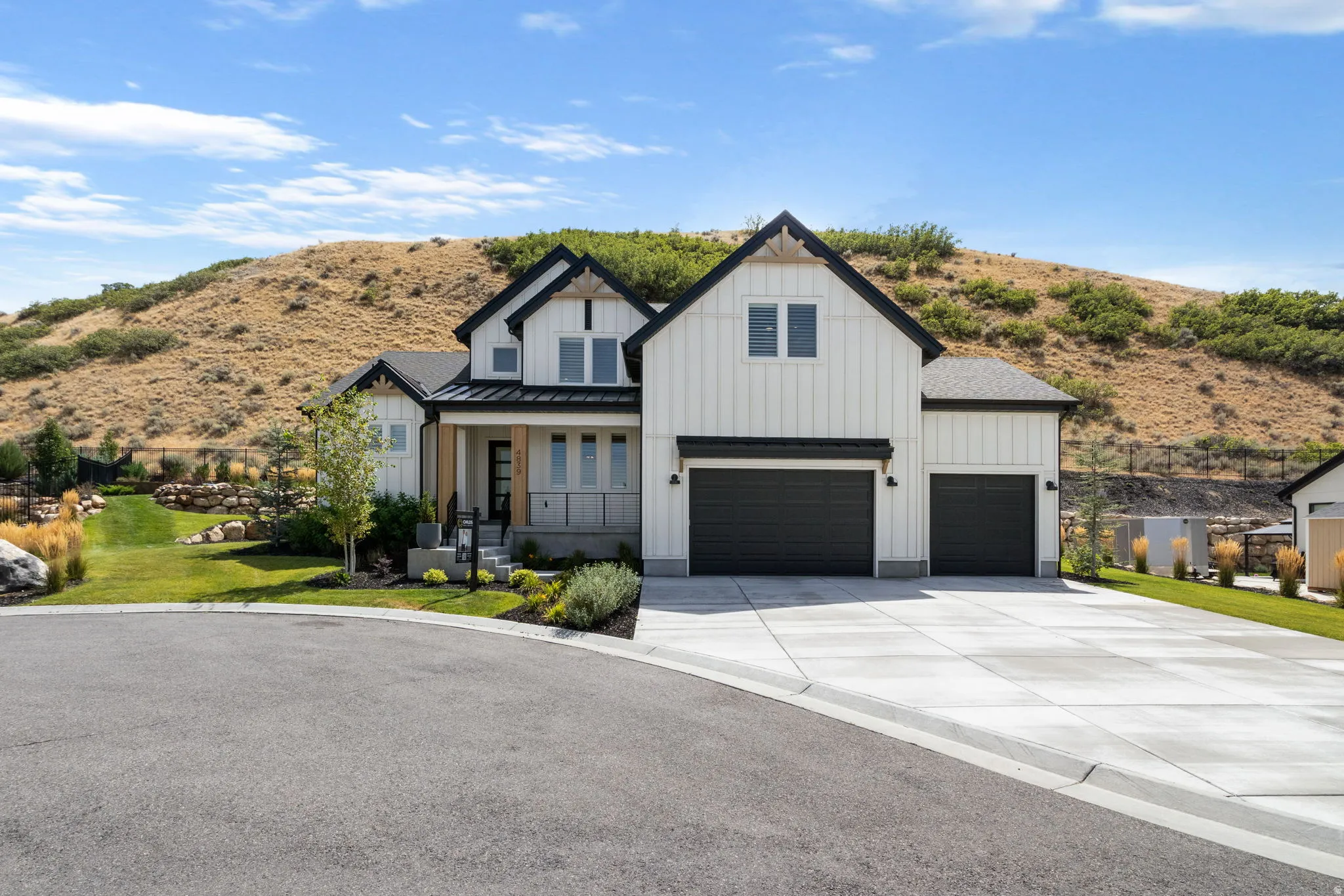White vertical siding exterior with contrasting black trim, covered front entry with natural wood pillars, two-car garage with black doors, concrete driveway, and front lawn with established landscaping