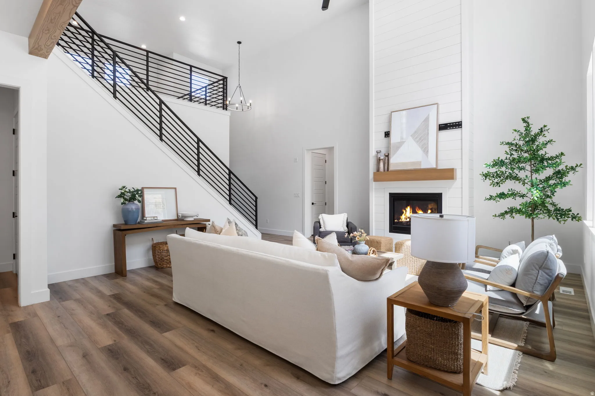 High ceilings with an exposed wood beam, wood-finish flooring, a contemporary fireplace with a shiplap surround and wood mantel, and a staircase featuring black horizontal iron railings