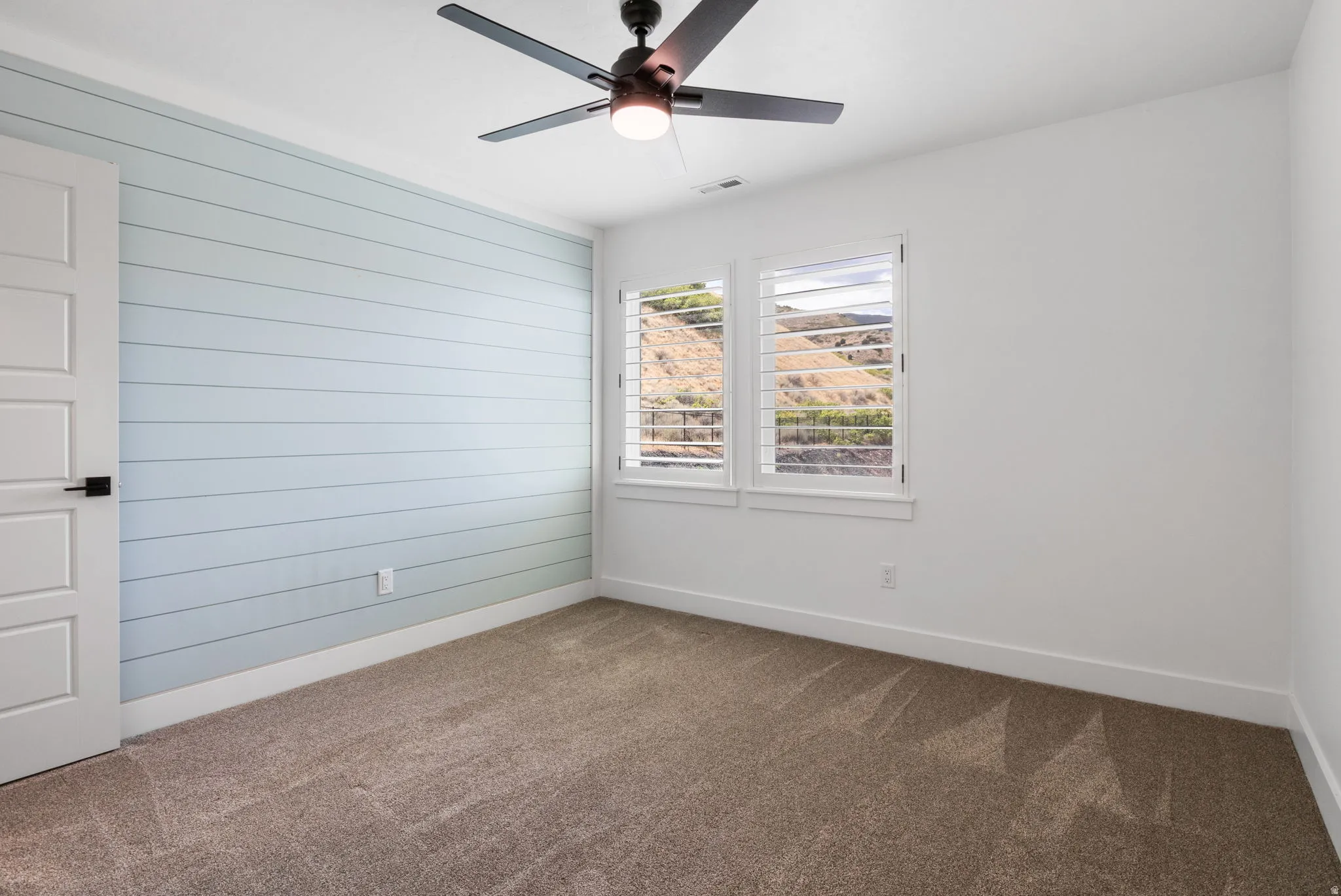 Light blue shiplap accent wall, two windows with white plantation shutters, white interior door with black hardware, ceiling fan with integrated lighting, and carpet flooring