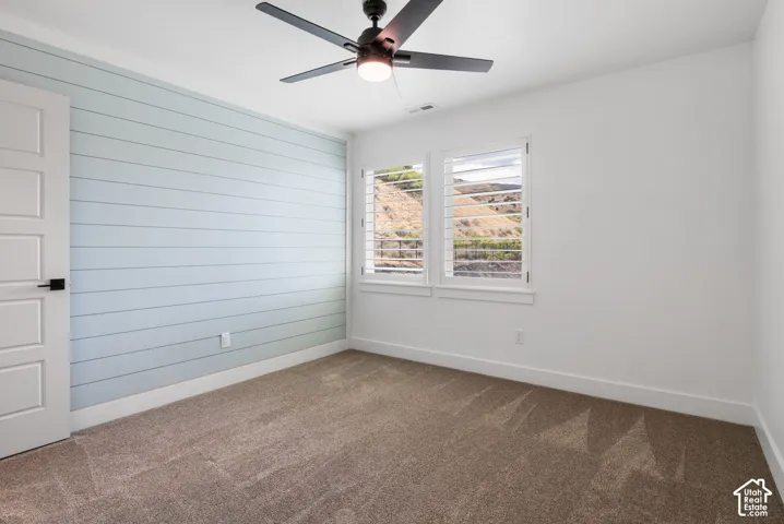 Light blue shiplap accent wall, two windows with white plantation shutters, white interior door with black hardware, ceiling fan with integrated lighting, and carpet flooring