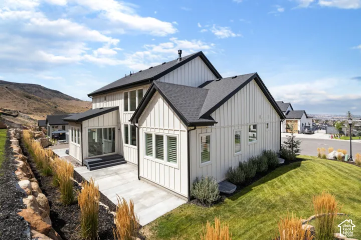 Board and batten siding, black trim accents, a dark shingled roof, a ground-level patio, and a tiered lawn