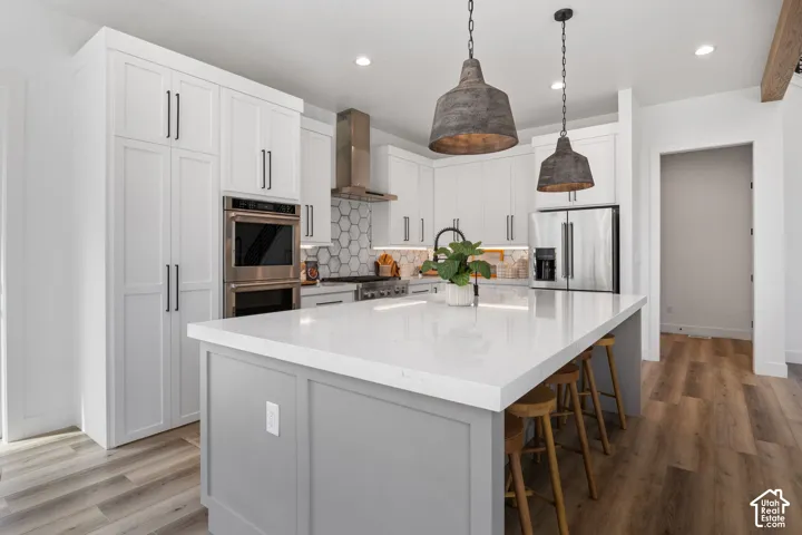 Kitchen featuring a central island with countertop seating, white shaker cabinetry, stainless steel appliances, a honeycomb tile backsplash, and wood-finish flooring