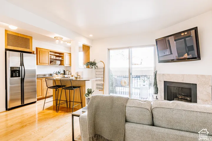 Light wood-finish flooring throughout, kitchen with stainless steel refrigerator, kitchen island with seating, fireplace with stone surround, and sliding glass door leading to a balcony