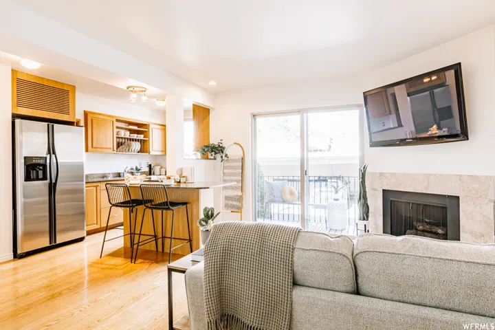 Light wood-finish flooring throughout, kitchen with stainless steel refrigerator, kitchen island with seating, fireplace with stone surround, and sliding glass door leading to a balcony