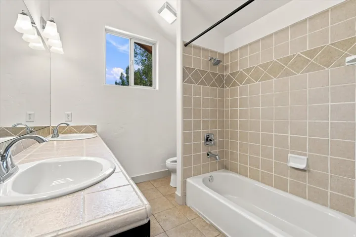 Dual vanity with integrated sinks, tiled countertop, large mirror, full-size bathtub, and tiled shower surround with decorative band