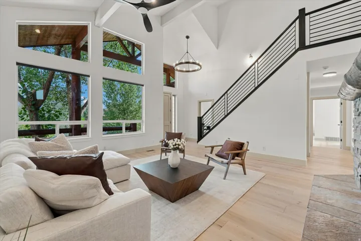 Vaulted ceiling with exposed beams, multiple large windows, light wood-finish flooring, linear black metal staircase railing, and a stone-clad fireplace