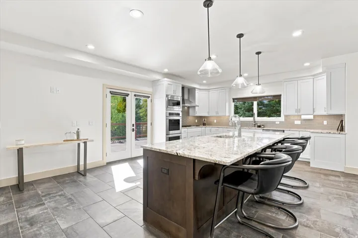 Kitchen featuring an expansive island with a light-toned countertop, built-in double ovens, white cabinetry, a neutral-toned tile backsplash, and dark-toned tile flooring
