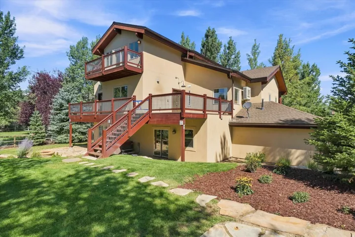 Multi-level decks with wood railing systems, ground-level patio access, stone slab pathway, mature landscaping, and stucco exterior