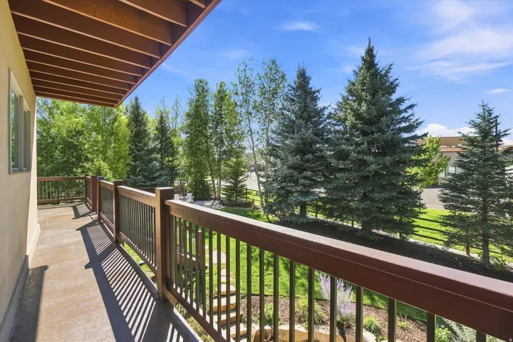 Covered balcony with wood railings and a concrete surface, overlooking mature trees and landscaped grounds