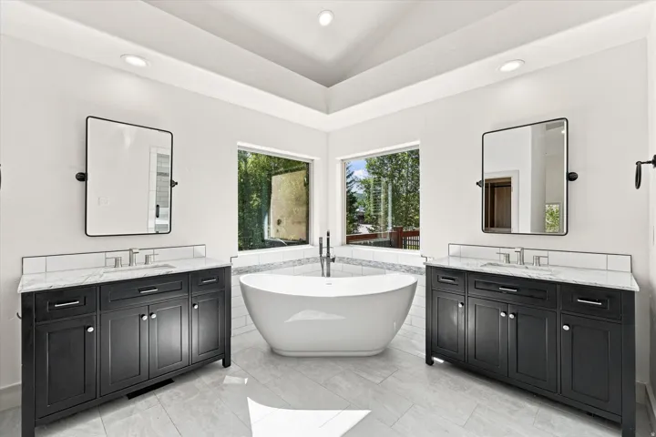 Freestanding soaking tub, dual vanity with undermount sinks, tile flooring, corner windows, and recessed lighting