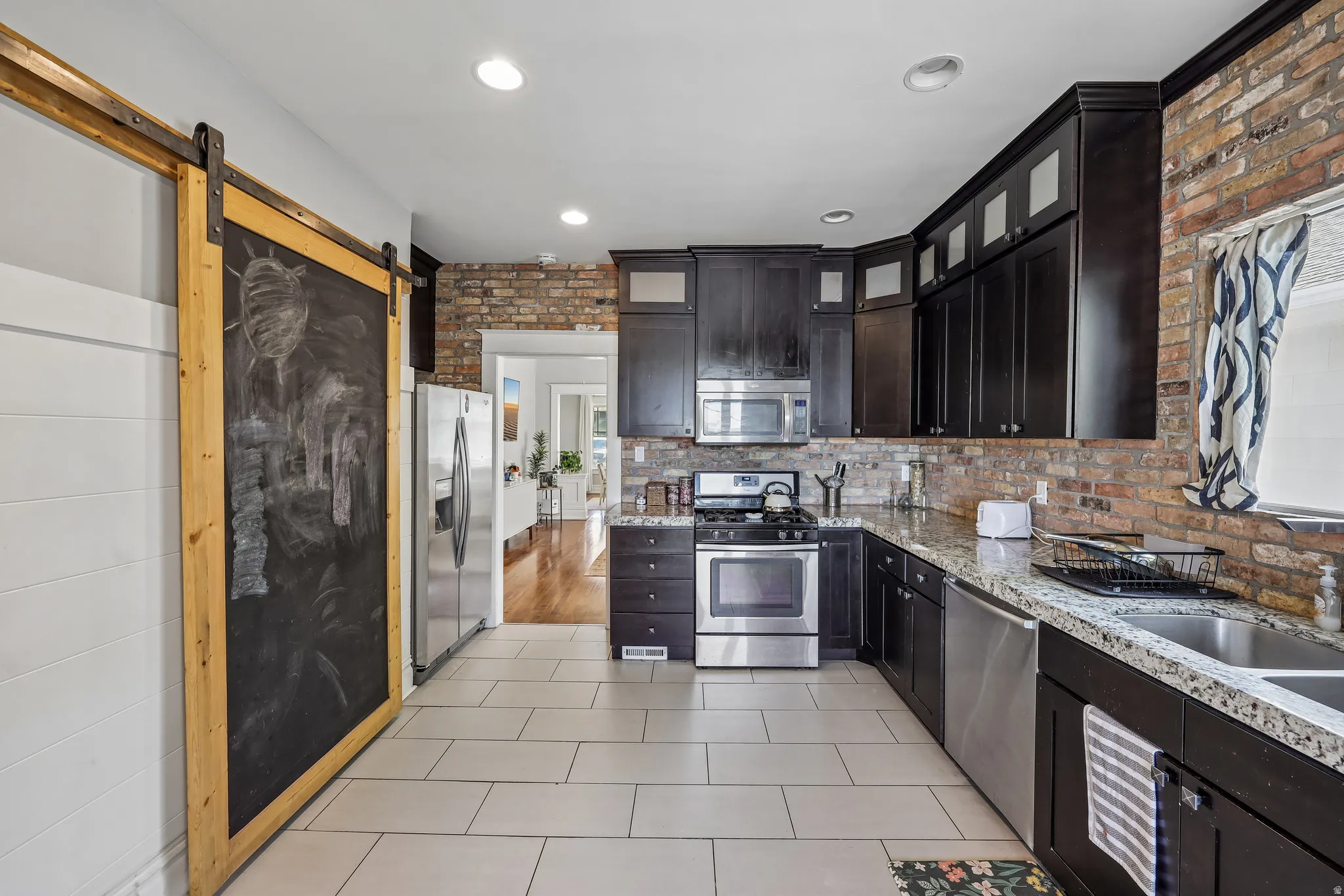 Kitchen featuring dark wood cabinetry, stainless steel appliances, brick accent wall, tile flooring, and a sliding barn door
