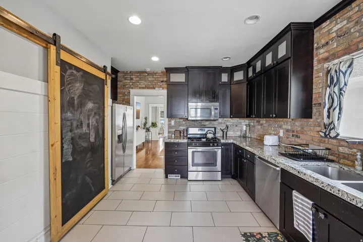 Kitchen featuring dark wood cabinetry, stainless steel appliances, brick accent wall, tile flooring, and a sliding barn door
