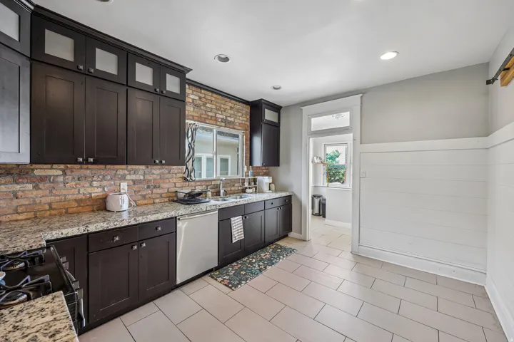 Kitchen featuring dark wood-finish cabinetry, granite countertops, brick-finish backsplash, stainless steel dishwasher, and recessed lighting