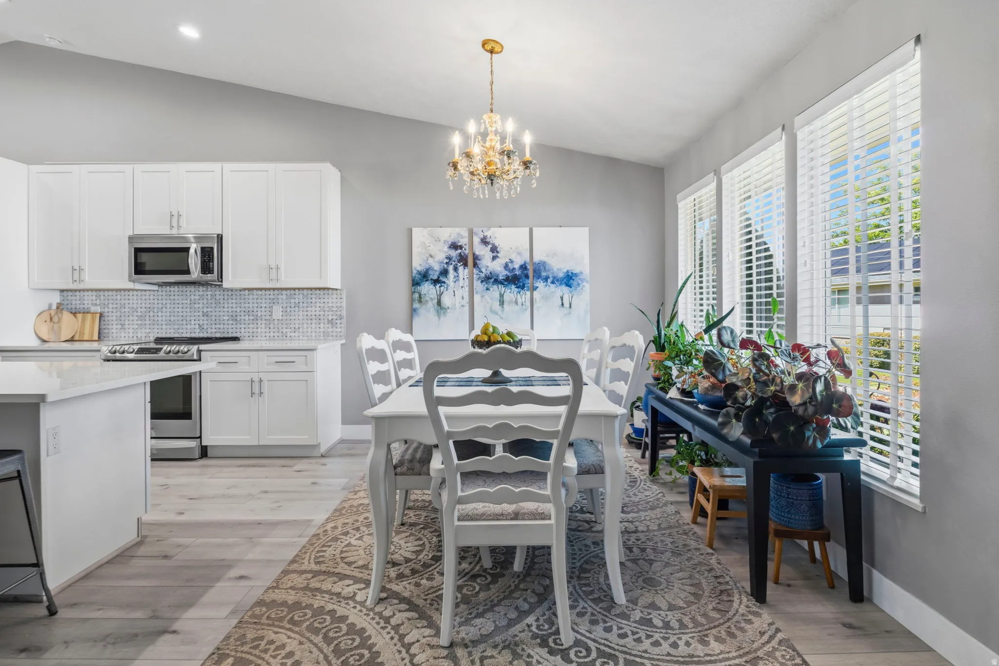 Vaulted ceiling with a chandelier, wood-finish flooring, white cabinetry, integrated stainless steel appliances, and multiple windows with blinds