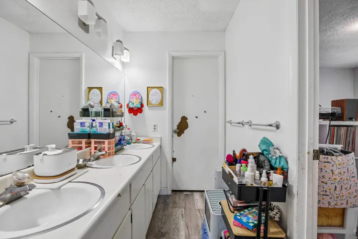 Dual vanity with individual sinks, expansive wall-mounted mirror, overhead vanity lighting, wood-finish flooring, and white cabinetry