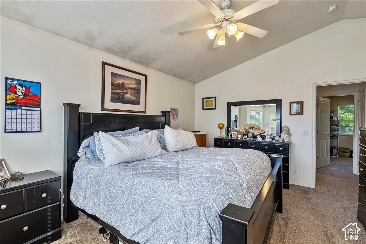 Vaulted ceiling, ceiling fan with integrated lighting, carpeted flooring, wall-mounted mirror, and an open doorway