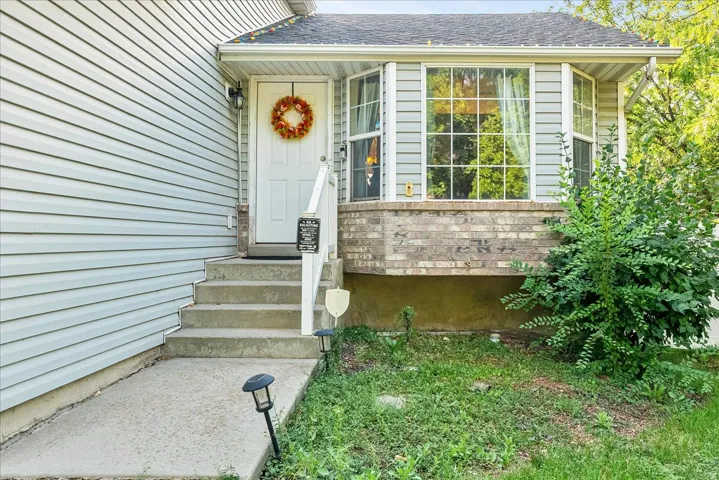 Light-toned siding exterior featuring a white entry door, concrete steps, and a bay window with a brick base