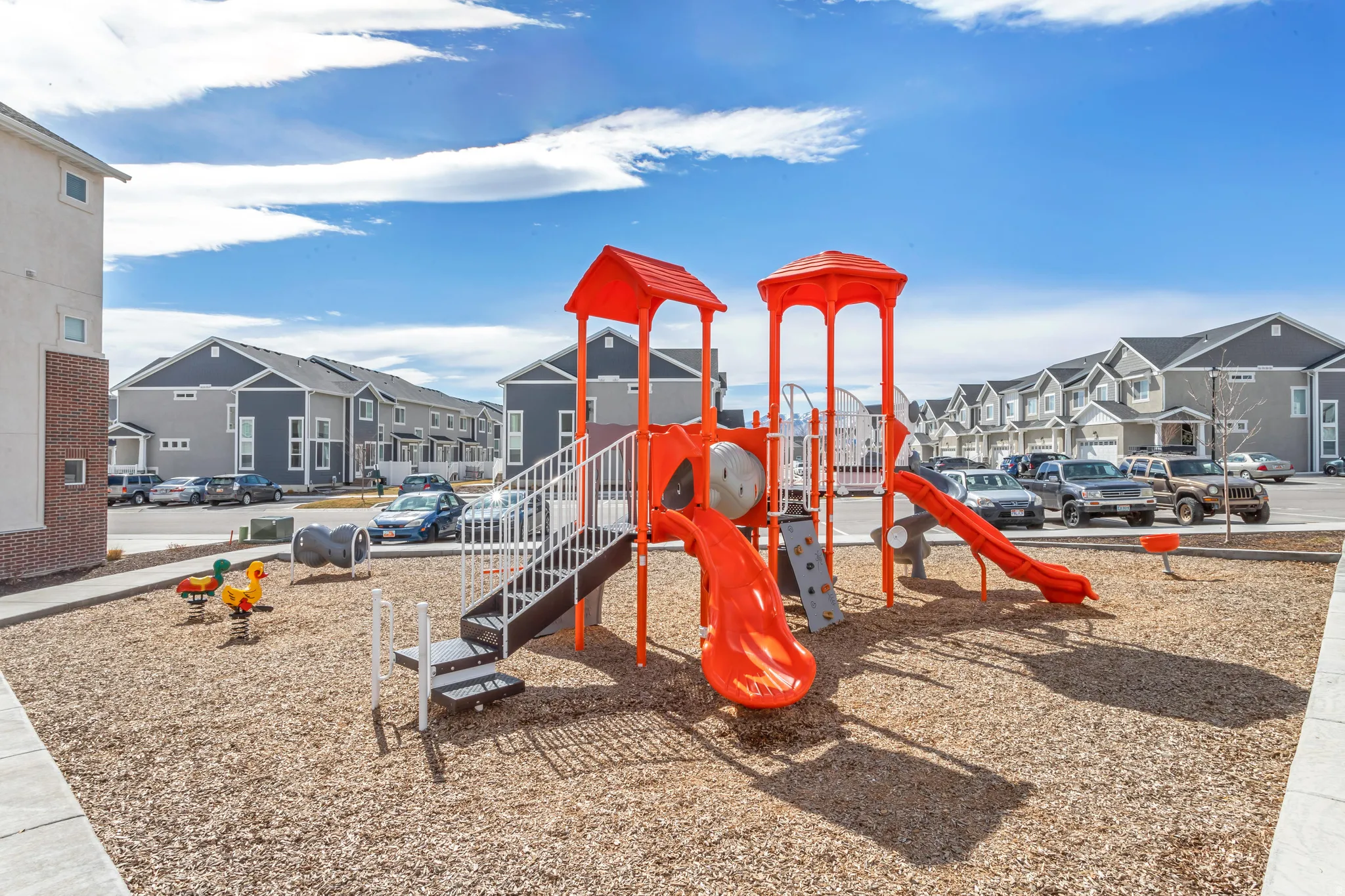 Recreational playground area featuring bright orange slides, climbing elements, and spring riders set on wood chip surfacing.