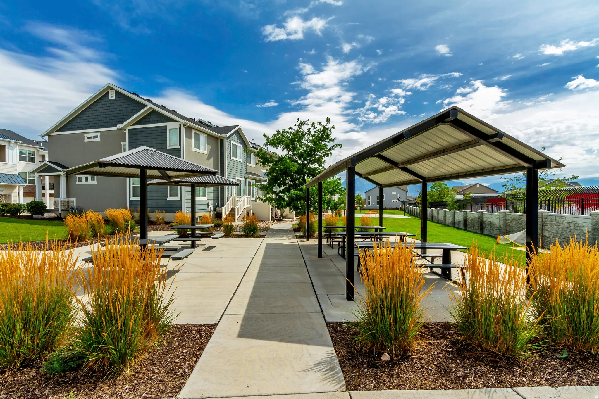 Community area featuring multiple covered picnic structures, concrete pathways, manicured lawn, and mature ornamental grasses.