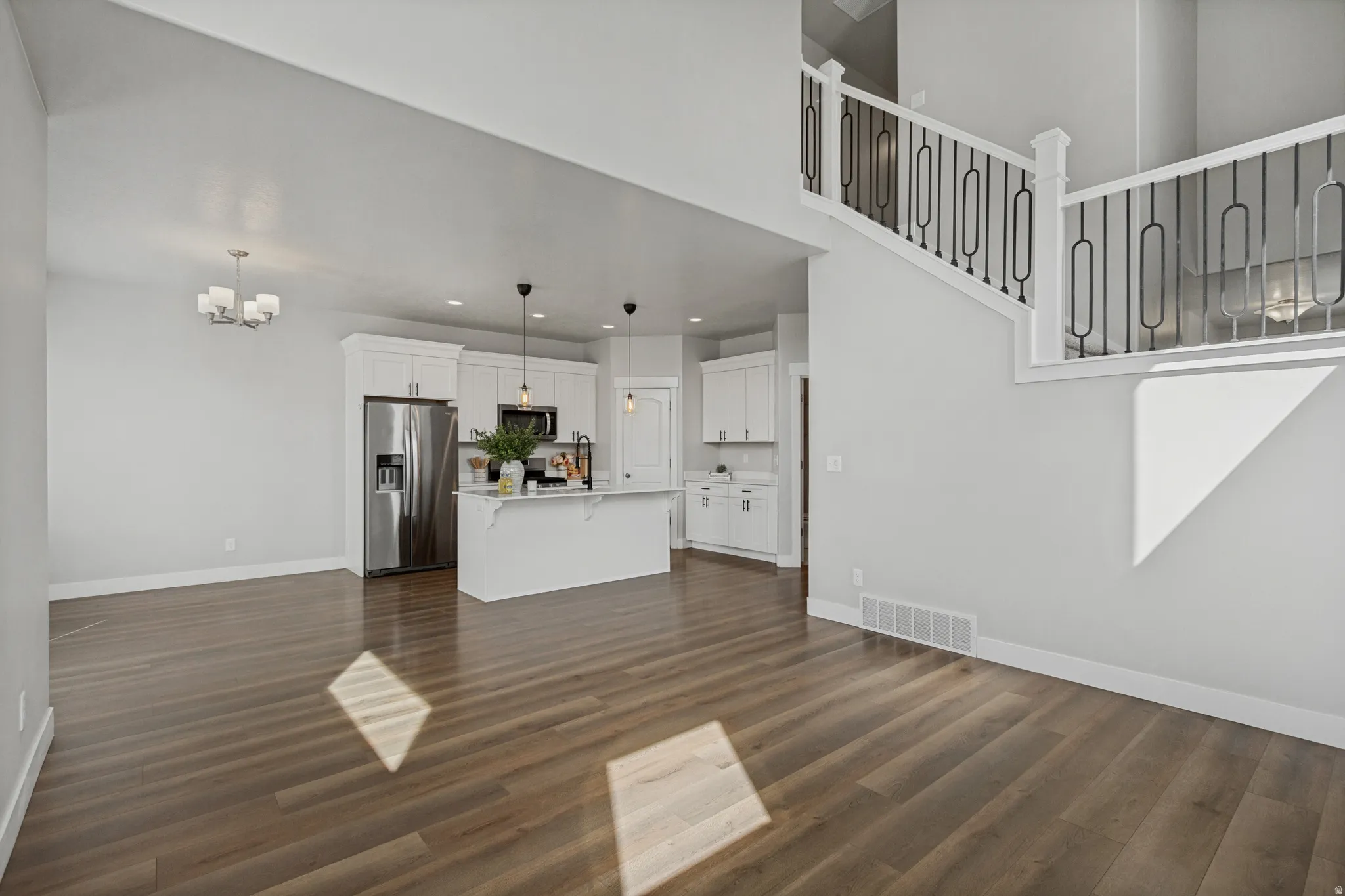 Expansive open-concept living area with wood-finish flooring, two-story ceiling, and a modern kitchen featuring white cabinetry, a large island with pendant lighting, and stainless steel appliances.