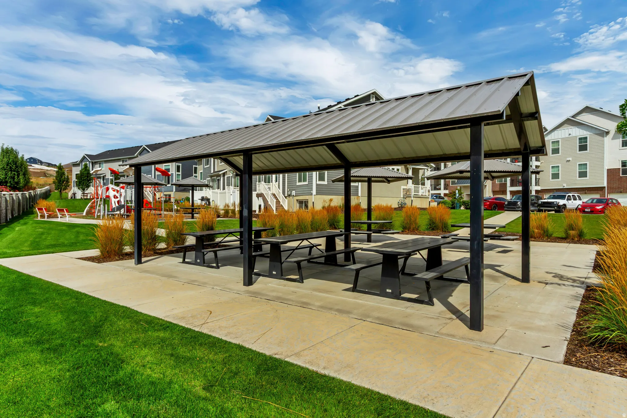 Covered outdoor area with multiple picnic tables, concrete hardscaping, and surrounding green lawn.