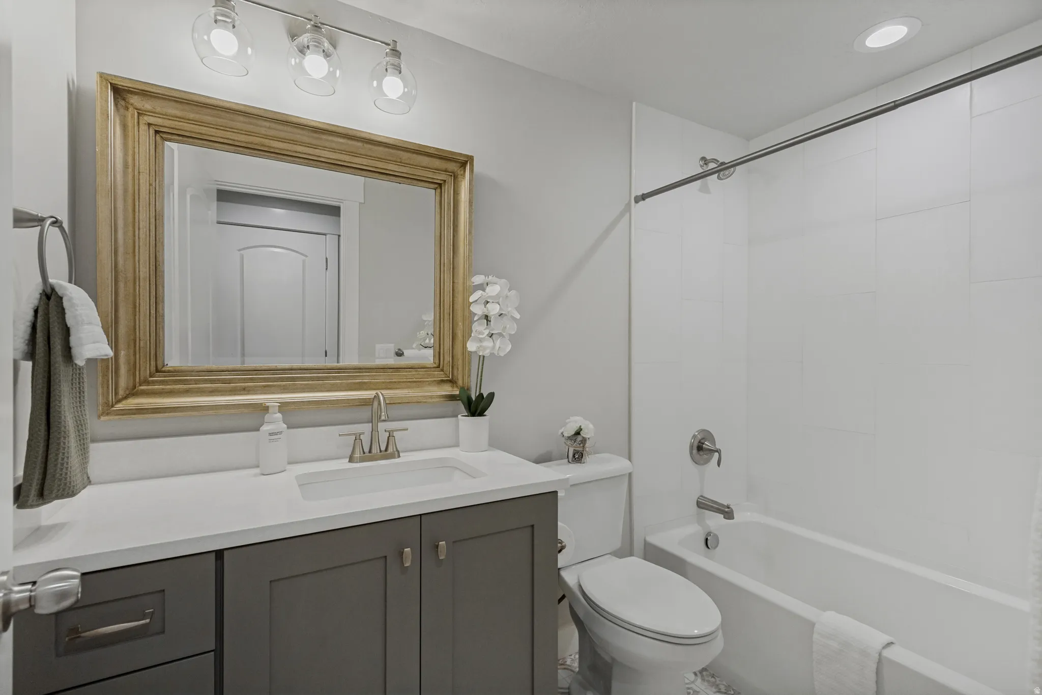 Vanity with a white countertop and gray cabinetry, undermount sink with a brushed nickel faucet, large framed mirror, and an adjacent white bathtub with a tiled surround.