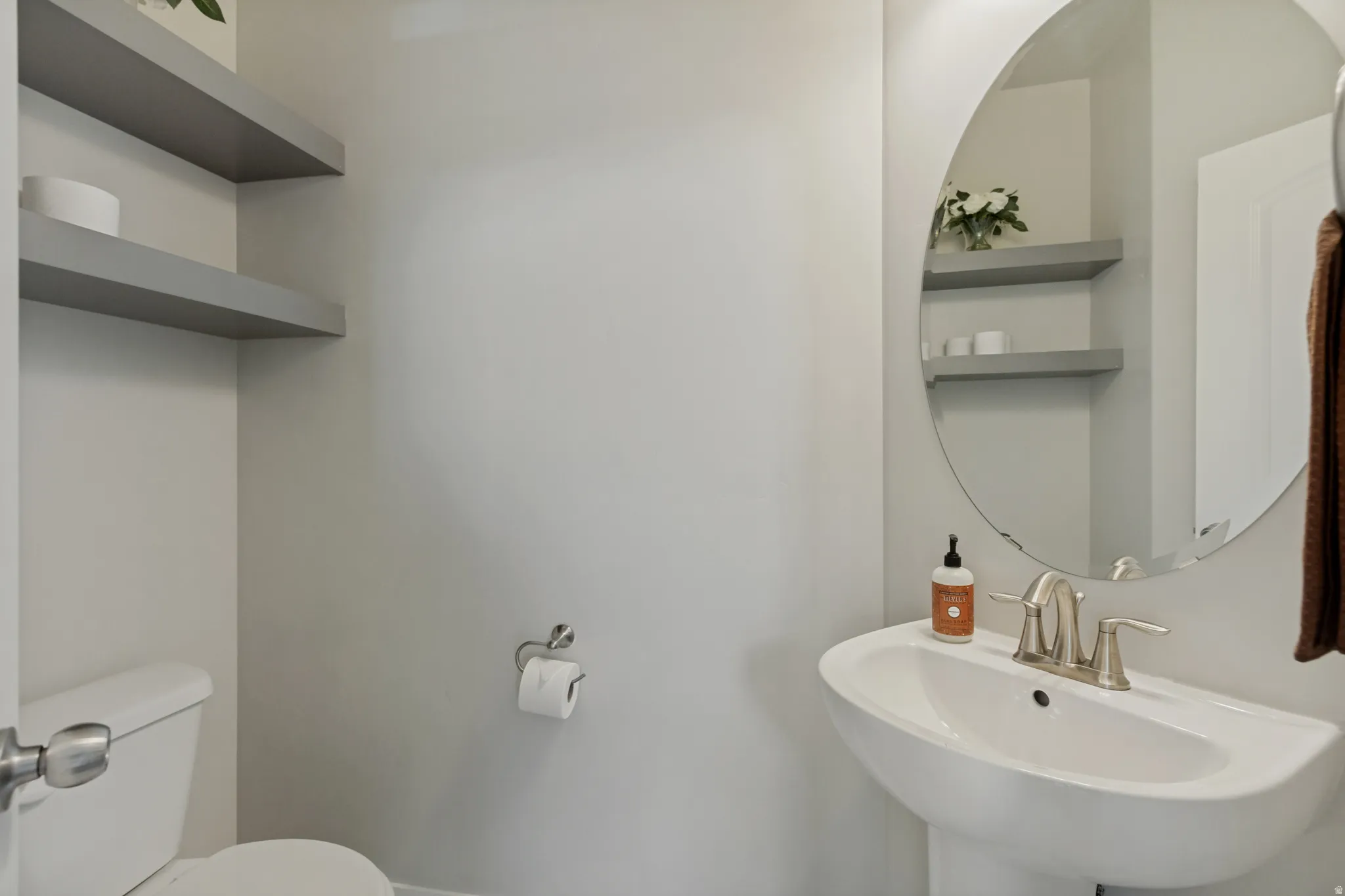 Half bathroom off the garage by kitchen. Pedestal sink with brushed nickel faucet, round frameless mirror, and built-in floating shelves.