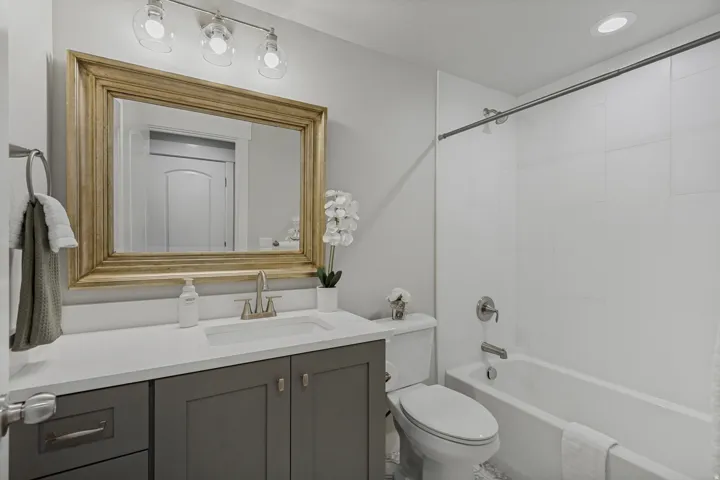 Vanity with a white countertop and gray cabinetry, undermount sink with a brushed nickel faucet, large framed mirror, and an adjacent white bathtub with a tiled surround.