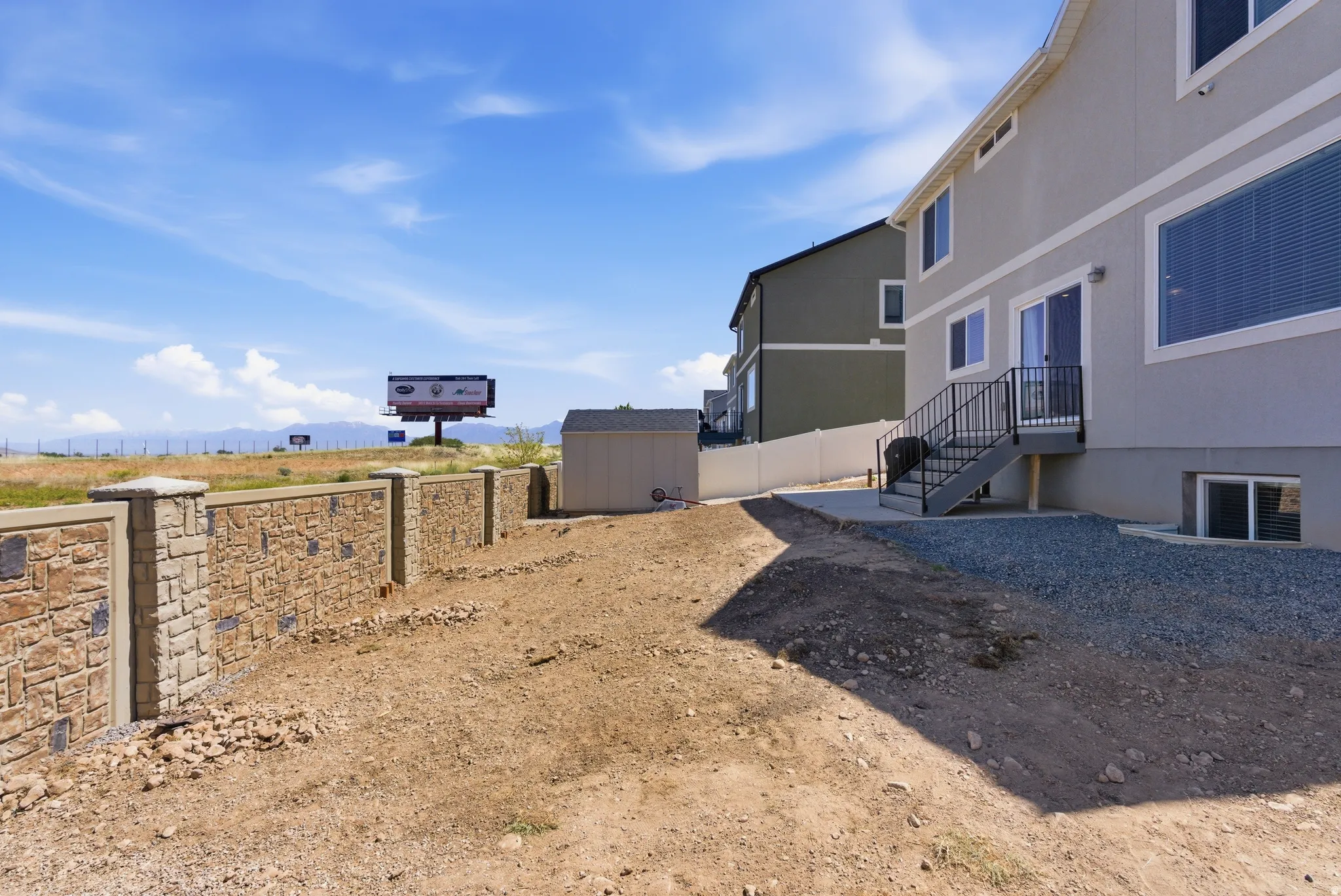 Stone-finish retaining wall, stucco exterior, sliding glass door, exterior stairs with metal railing, and a detached shed