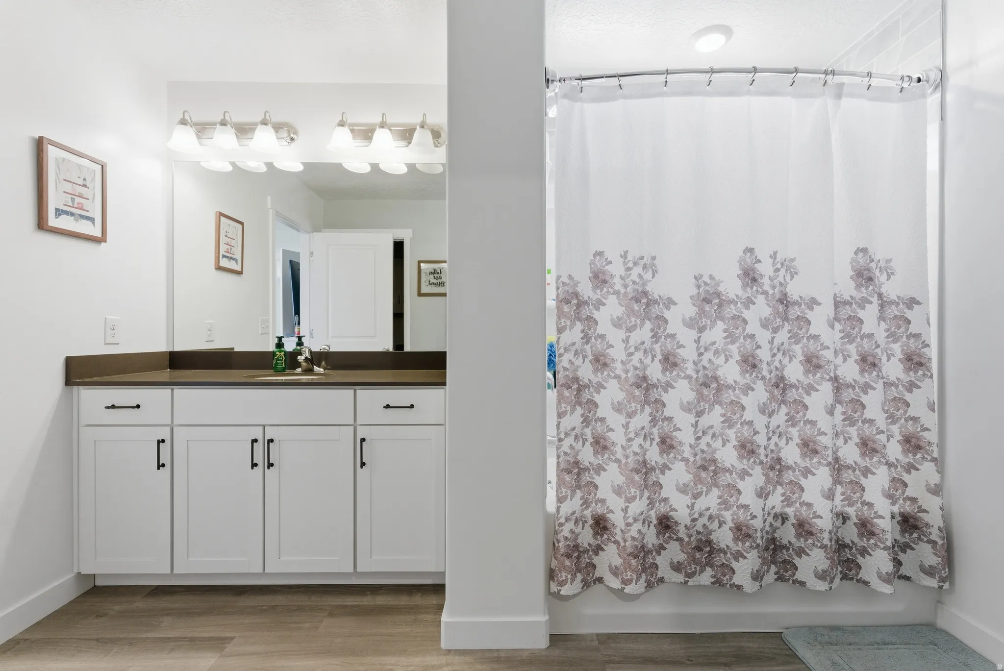 White shaker cabinetry with matte black hardware, dark countertop, full-width vanity mirror, and wood-finish flooring