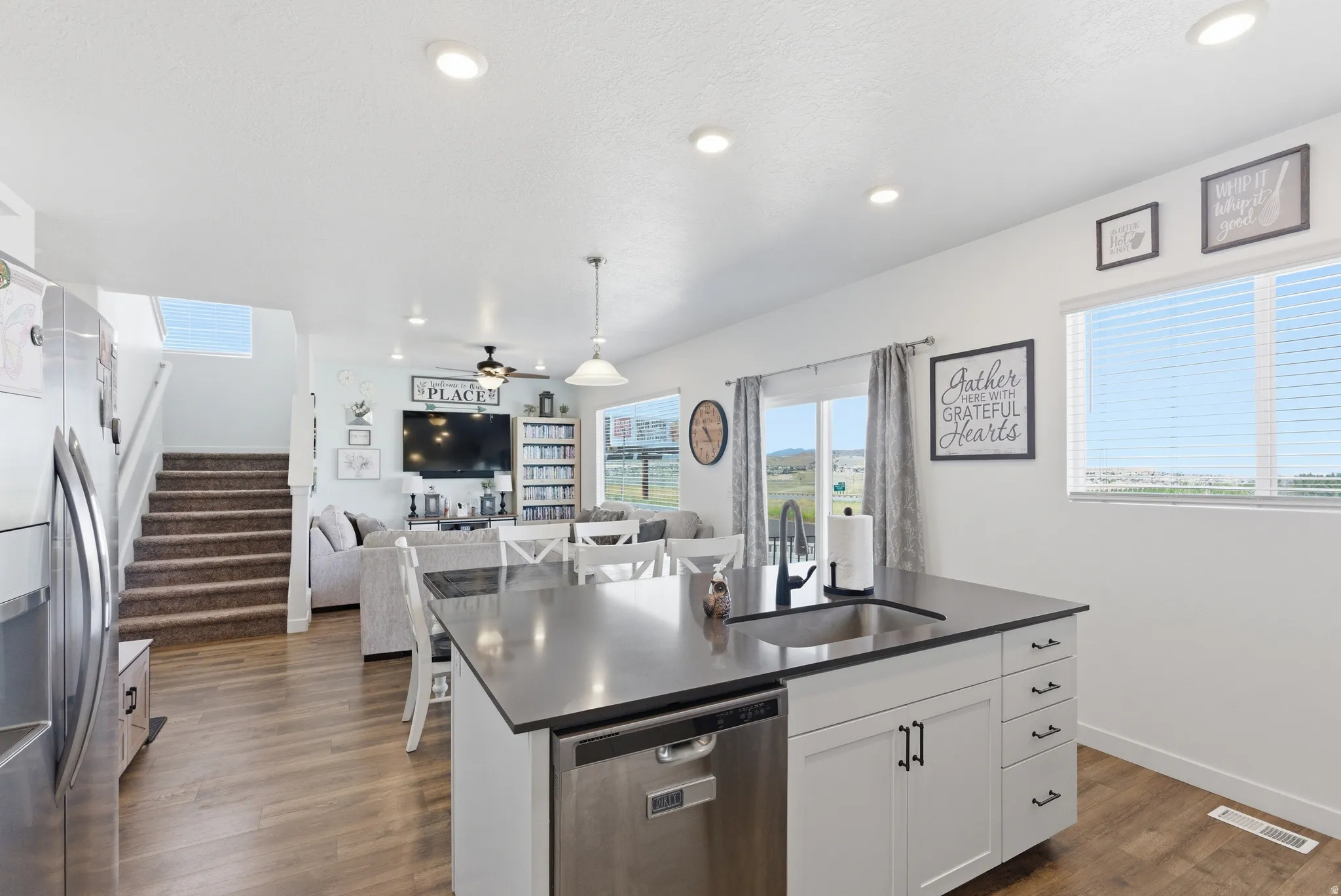 Open-concept living space featuring wood-finish flooring, recessed lighting, a kitchen island with an undermount sink, and a staircase with carpeted treads